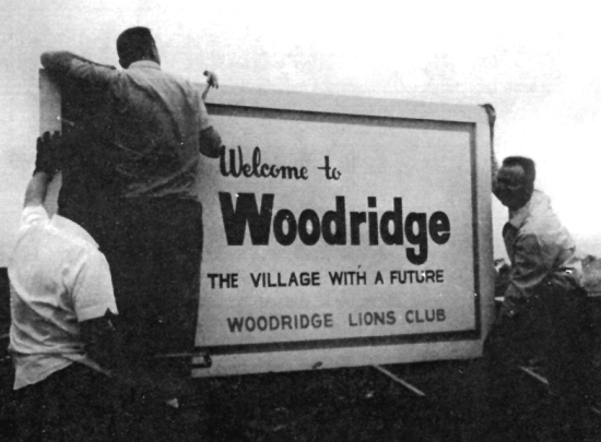 Historical black and white image of three men putting up a "Welcome to Woodridge" sign: "The Village with a Future; Woodridge Lions Club"