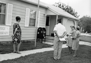 The Library as it was when it first opened in 1969. It is inside of a single-level house with light-colored siding. Part of the Woodridge Public Library sign is visible. Four people are standing outside the library.