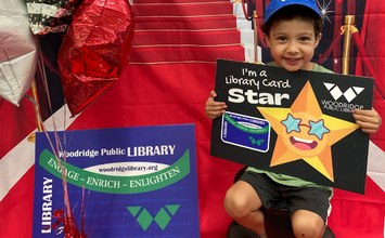 Child on a red carpet, holding a library card sign next to a large library card.
