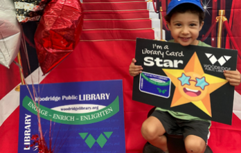 Child on a red carpet, holding a library card sign next to a large library card.