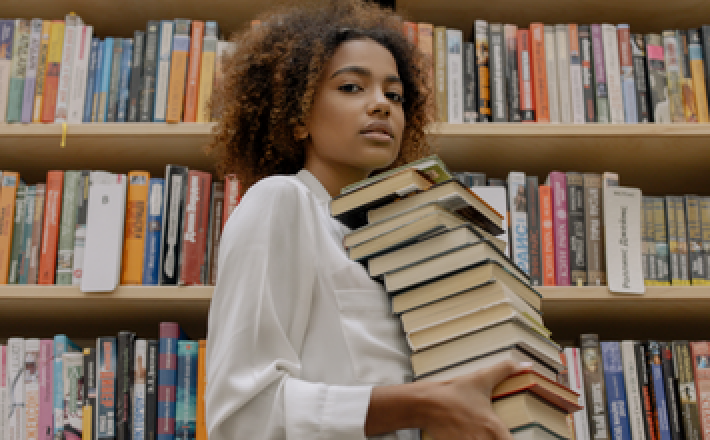 Image of a Black woman with curly hair in front of a book shelf and holding a stack of books